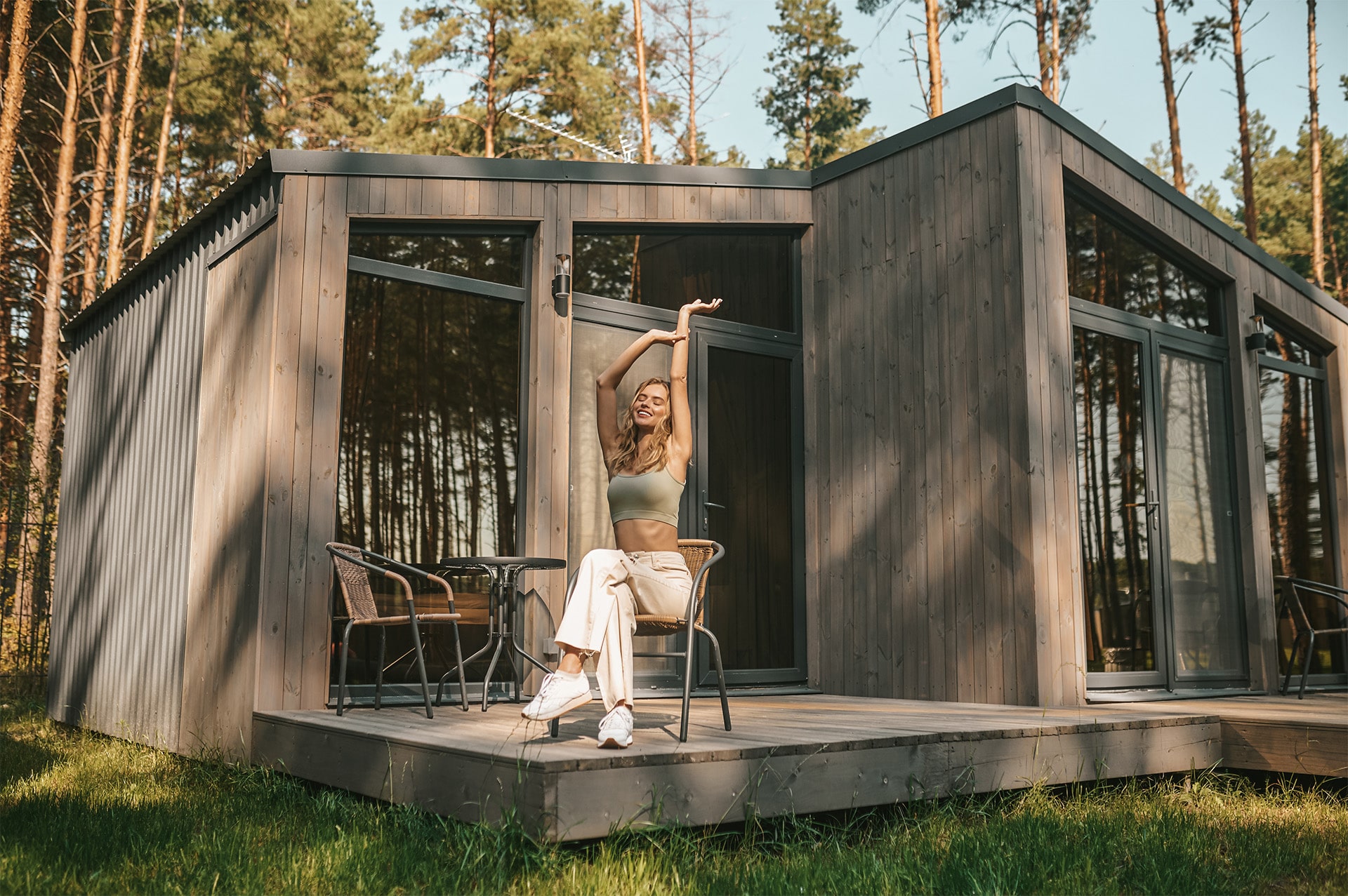Glückliche Blondine freut sich auf Terrasse über neues Tiny Home aus Holz in modularer Bauweise