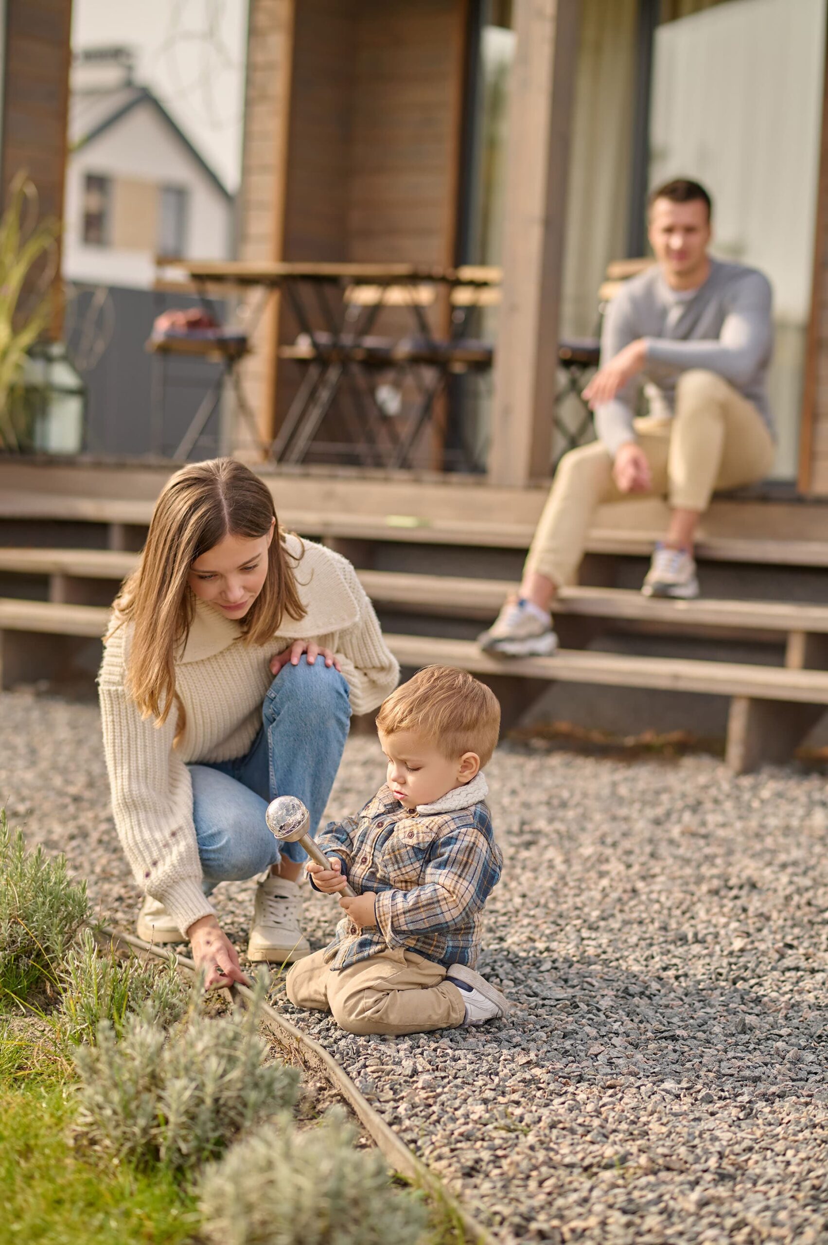 Jungfamilie vor ihrem Tiny Home, Mutter spielt mit Kleinkind in Wiese während der Vater auf der Veranda sitzt.