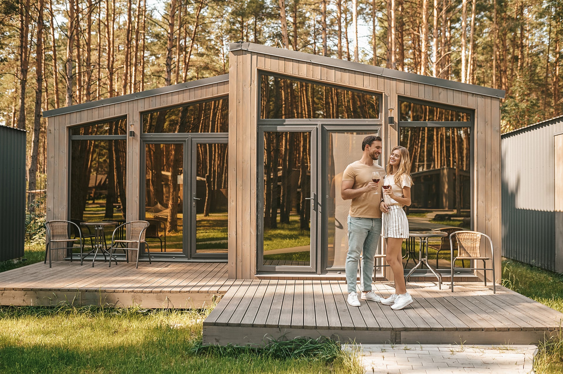 Verliebtes Pärchen steht auf Terrasse eines modularem Holzbau Hauses und trinkt Wein.